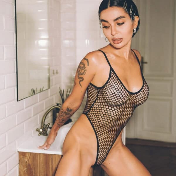 Black bodysuit in fine Pivovarova fishnet, scalloped cut and thin straps, worn on a feminine silhouette in an interior, selection Brigade Mondaine Paris.