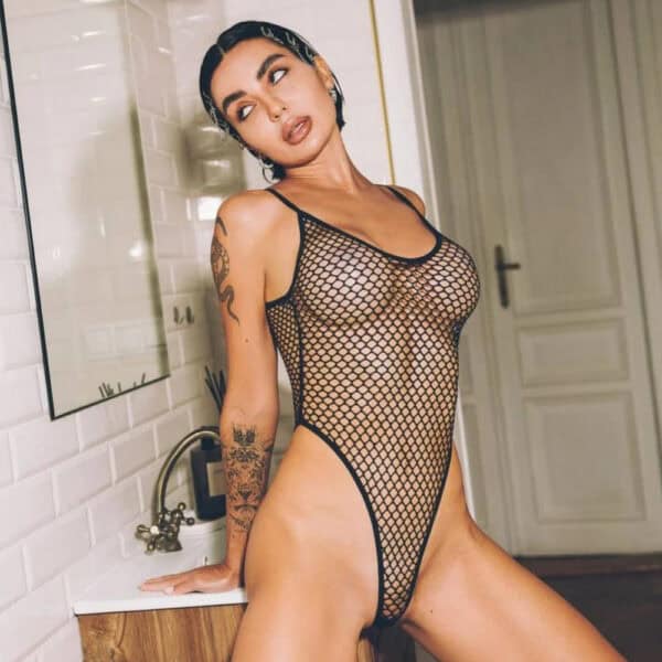 Black bodysuit in fine Pivovarova fishnet, scalloped cut and thin straps, worn on a feminine silhouette in an interior, selection Brigade Mondaine Paris.
